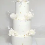 עוגת חתונה מעוצבת בשלוש קומות, מקושטת בפרחים לבנים ועלי זהב. מתאימה לאירועים מיוחדים.