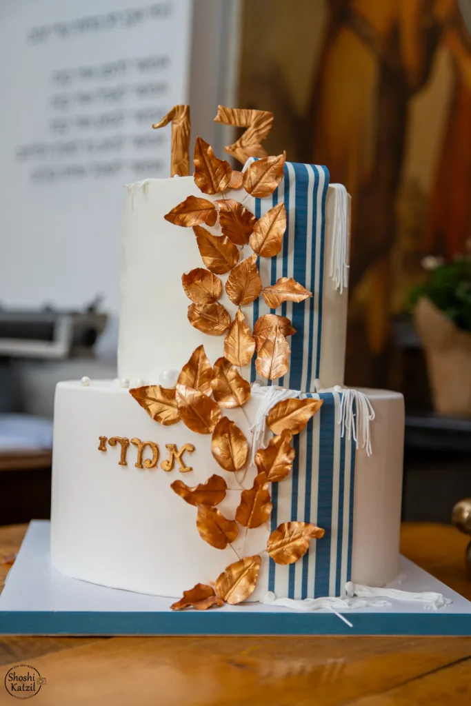 עוגת בר מצווה עם עלי זהב אכילים כקישוט ופסים כחול לבן של טלית על העוגה הלבנה